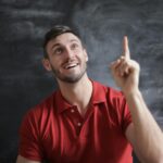 Happy young man in a red polo shirt smiling and pointing upwards in front of a blackboard.