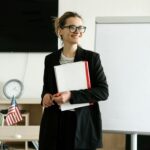 Smiling woman in black blazer holding notebooks next to a whiteboard with a US flag in an office setting.