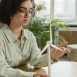 Focused young woman studying a wind turbine model indoors, highlighting renewable energy and education.
